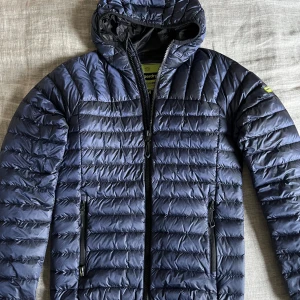 Mörkblå dunjacka från Superdry med huva - Mörkblå dunjacka från Superdry med huva och snygg quiltad design. Jackan har dragkedja framtill, två sidofickor med dragkedja och en cool Superdry-logga på ärmen. Perfekt för kalla dagar och har en modern, sportig look.