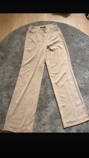 Beige wide kostymbyxor med dragkedja - Snygga beige kostymbyxor från Bikbok med wide fit och hög midja. Byxorna har en diskret dragkedja i sidan och är tillverkade i ett mjukt, slätt material som ger en clean look. Perfekta för dig som gillar stilrena och trendiga plagg.