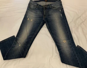 Blå Luke jeans från Lee W31 L34 - Snygga blå jeans från Lee, modell Luke, med klassisk femficksdesign och slitna detaljer på benen. Jeansen har en rak passform och är tillverkade i slitstarkt denimtyg. Perfekt för dig som gillar en cool och avslappnad stil.