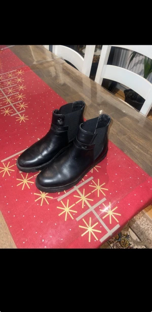 Svarta chelsea boots i skinn - Säljer ett par svarta chelsea boots med rund tå och platt sula. Bootsens ovandel är i slätt skinn och har elastiska paneler på sidorna för enkel påtagning. Yttersulan är grovmönstrad för extra grepp. Perfekt för dig som gillar stilrena och robusta skor.