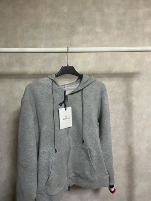 Grå Moncler hoodie med dragkedja - Snygg grå hoodie från Moncler med dragkedja och huva. Tröjan har snörning i huvan, två fickor fram och Moncler-logga på ärmen. På huvan finns coola ränder i rött, vitt och mörkblått. Tillverkad i mjuk bomull, perfekt för chill dagar.