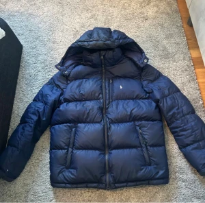 Mörkblå pufferjacka från Polo Ralph Lauren - Säljer en fet mörkblå pufferjacka från Polo Ralph Lauren med huva och dragkedja framtill. Jackan har två sidofickor med dragkedja och en bröstficka, samt den klassiska Polo-loggan broderad i guld på bröstet. Perfekt för kalla dagar och riktigt snygg streetstil.