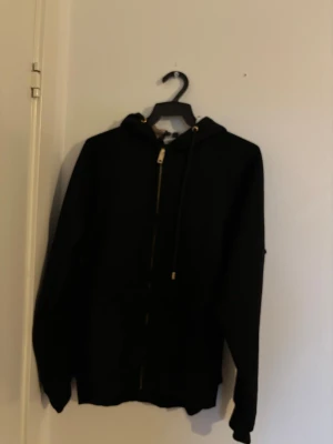 Svart hoodie med Burberry-rutig huva - Svart hoodie med dragkedja och snörning, samt en huva fodrad med klassiskt Burberry-rutigt tyg i beige, rött och svart. Långärmad och med ribbade muddar. Perfekt för dig som vill ha en clean look med en lyxig twist.