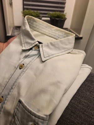 Vit denimskjorta från Bershka - Vit denimskjorta från Bershka med kontrasterande svarta sömmar, två bröstfickor med lock och knappar i trälook. Skjortan har klassisk krage och långa ärmar med knapp vid manschetten. Perfekt för en cool och avslappnad stil.