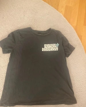 Gina tricot - Cool svart t-shirt med unikt grafiskt tryck i vitt och ljusblått både fram och bak. Trycket har en vågig, rutig design och texten 'Radiate Positivity' med blommor. T-shirten har rund hals och korta ärmar, perfekt för dig som gillar statement-plagg.