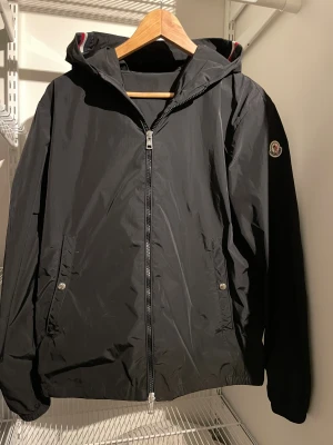 Svart vindjacka från Moncler - Svart vindjacka från Moncler med huva och dragkedja framtill. Jackan har Moncler-logga på ärmen och coola ränder i rött, vitt och blått på insidan av huvan. Två fickor med tryckknappar och lätt, smidig polyester som passar perfekt till Grisch stilen.