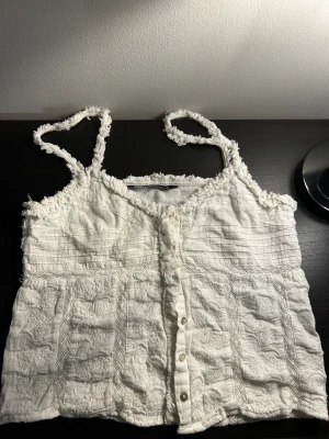 Vit linnetopp med knappar från Zara - Supersöt vit linnetopp från Zara med smala axelband och dekorativa fransdetaljer. Toppen har knäppning framtill och ett strukturerat tyg med broderade partier. Perfekt för en somrig look och lätt att matcha med jeans eller kjol.
