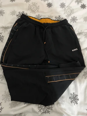 Svarta mjukisbyxor från BOSS - Svarta mjukisbyxor från BOSS med orange detaljer och logotyp längs sidorna. Byxorna har snörning i midjan och en bekväm passform. Insidan är fodrad med orange tyg för extra stilpoäng. Perfekta för chill eller streetwear.