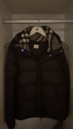 Svart Burberry pufferjacka med huva - Svart pufferjacka från Burberry med klassisk rutig huva och tydlig Burberry patch på ärmen. Jackan har dragkedja, snörning vid huvan och quiltad design. Perfekt för kalla dagar och har en riktigt snygg streetstil. Tar gärna emot byten (nrkp)