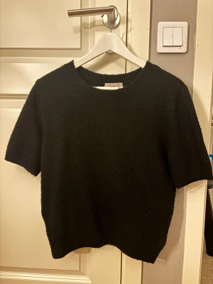 Svart stickad tröja  - En svart stickad tröja från H&M med korta ärmar och rund halsringning. Tröjan har en enkel och clean look, perfekt för lager-på-lager eller att bära som den är. Mjuk och skön känsla tack vare det stickade materialet.