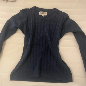 Marinblå stickad tröja Blue Leage - Snygg marinblå stickad tröja från Blue Leage Equestrian med v-ringad hals och flätstickat mönster framtill. Tröjan har långärmad passform och sitter tight på kroppen, perfekt till jeans eller kjol. Ribbad kant vid hals och ärmslut.