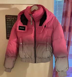 Beshka rosa-grå pufferjacka med huva - Säljer en fett snygg pufferjacka från Bershka i rosa och grått med cool ombre-effekt. Jackan har stor huva, dragkedja framtill, två fickor med knappar och ribbade muddar. Reflekterande material som syns i mörker, perfekt för vinter och höst. Croppad passform.
