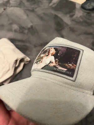 MAGGIORE keps med retrotryck - Snygg ljusgrå keps från MAGGIORE med mesh baktill och justerbar snapback. Framtill finns ett retroinspirerat tryck med ikonisk bild. Framsidan är i mjuk mockaliknande material och baksidan i grå mesh för extra ventilation.