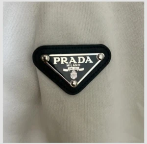 Vit långärmad tröja från Prada - Vit långärmad tröja från Prada med klassisk svart och silver logotyp på bröstet. Tröjan har rund hals och är tillverkad i mjukt bomullsmaterial. Enkel och clean design som ger en lyxig vibe, perfekt för dig som gillar stilrena plagg med exklusiv touch.