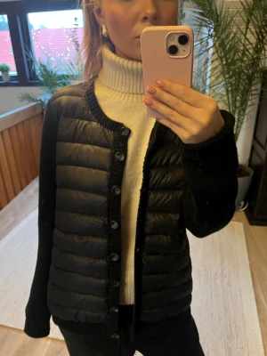 Svart cardigan  - Svart cardigan  med stickade ärmar och ribbad krage. Jackan har quiltad front i glansig polyester och knäpps med knappar. Snygg, stilren och perfekt för dig som gillar en modern look med coola detaljer.