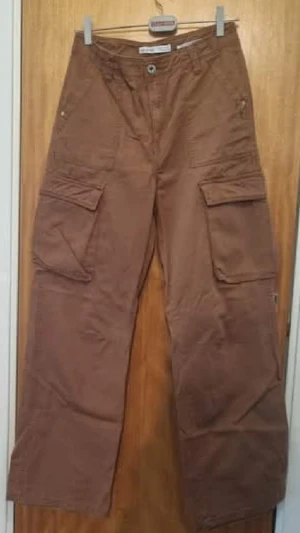 Bruna cargopants med stora fickor - Säljer ett par bruna cargopants med bred passform och flera stora fickor på benen. Byxorna har klassisk midja och är tillverkade i ett slitstarkt bomullsmaterial. Perfekta för dig som gillar streetwear och vill ha mycket plats för prylar.