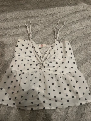 Prickig vit linnetopp med volang - Supersöt vit linnetopp med svarta prickar, smala axelband och twistad v-ringning framtill. Toppen har volang nedtill och scrunchad där fram. Säljer för att den är för liten