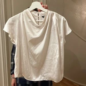 Vit sidenblus från Zara med drapering - Säljer en vit blus från Zara i mjukt sidenmaterial med snygg drapering framtill. Blusen har korta ärmar och knappar i ryggen för en clean look. Perfekt för dig som gillar stilrena och eleganta toppar med lite extra detalj. Knappt använd men inte struken så därför ser den mer sliten ut på bild.