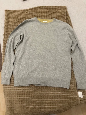 Grå stickad tröja Tom Tailor - Snygg grå stickad tröja från Tom Tailor med rund hals och diskret gul detalj på insidan av kragen. Tröjan har långärmade och ribbade muddar vid ärmslut och nederkant. Perfekt för dig som gillar enkel och stilren design.