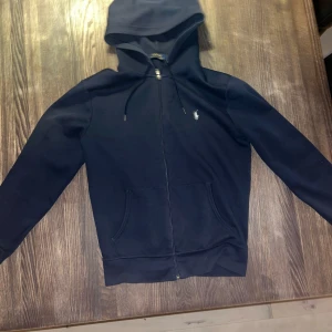 Marinblå hoodie från Polo Ralph Lauren - Snygg marinblå hoodie från Polo Ralph Lauren med klassisk logga broderad i vitt på bröstet. Tröjan har huva med snörning, dragkedja framtill och två fickor. Tillverkad i mjuk bomull, perfekt för chill dagar. Passformen är normal och ärmarna är långa. De ända minus med den är att den har tre små hål längst upp brevid dragkedjan men det syns inte ifall man har den half zip o vikt.