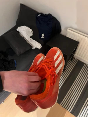 Adidas röda fotbollsskor med dobbar - Säljer ett par röda Adidas fotbollsskor med vita ränder och snörning. Skorna har en låg profil och är utrustade med gula och vita dobbar på sulan för grymt grepp på planen. Ovandelen är i syntetmaterial och designen är sportig med rund tå. Köpte för 1200kr förra året och har inte använt mycket alls 