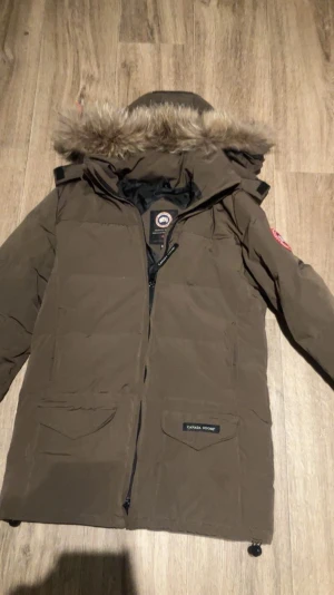 Brun Canada Goose dunjacka med päls - Säljer en äkta brun dunjacka från Canada Goose med äkta päls på huvan, klassisk logga på ärmen och två stora fickor framtill. Jackan har lång ärm, dragkedja och är fodrad med svart insida. Perfekt för kalla dagar. Pris kan diskuteras vid snabb affär