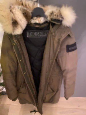 Beige parkajacka med päls från The Smirk - Parkajacka från The Smirk i beige med fluffig päls på huvan. Yttertyg i bomull och fodrad med polyester för extra värme. Jackan har dragkedja, knappar och ribbade muddar vid ärmslut. Perfekt för kalla dagar och har snygg urban vibe.