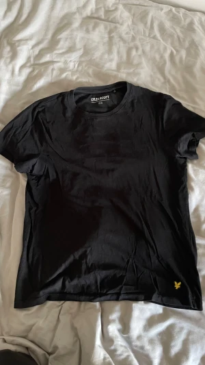 Svart t-shirt från Lyle & Scott - Svart t-shirt från Lyle & Scott med klassisk rund hals och korta ärmar. T-shirten har en liten gul broderad logga nere vid höger fåll. Mjuk och skön bomull som passar perfekt till jeans eller shorts. Enkel och stilren design.