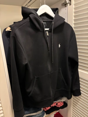 Svart hoodie från Polo Ralph Lauren - Svart hoodie från Polo Ralph Lauren med klassisk logga på bröstet. Tröjan har huva med snörning, dragkedja framtill och två fickor. Tillverkad i mjuk bomull och har långärmad passform. Perfekt för chill dagar och enkel att matcha med jeans eller joggers.
