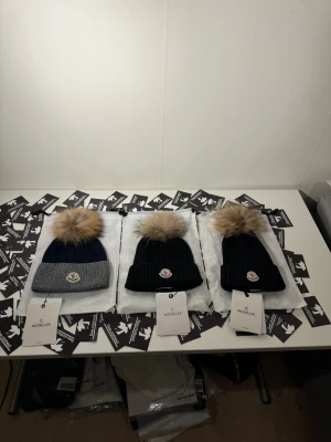 Moncler mössa med pälsboll - Snygg ribbstickad mössa från Moncler med stor beige pälsboll på toppen och klassisk logga framtill. Finns i svart och mörkblått med grå uppvikt kant. Perfekt för dig som vill ha en lyxig och trendig vinteraccessoar.