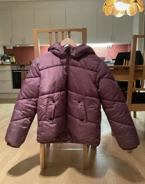Lila pufferjacka från H&M med huva - Säljer en lila pufferjacka från H&M i storlek 152. Jackan har en stor och mysig huva, två stora fickor framtill med knappar och dragkedja. Perfekt för kalla dagar när du vill hålla dig varm och ändå se snygg ut.