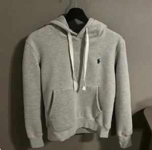 Grå hoodie från Polo Ralph Lauren - Snygg ljusgrå hoodie från Polo Ralph Lauren med klassisk broderad logga på bröstet. Tröjan har huva med vita snören, känguruficka och ribbade muddar. Mjuk och skön i bomullsmix, perfekt för chill dagar. Custom fit-modell för en normal passform.