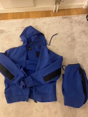 Blå hoodie från BOSS med svarta detaljer - Säljer en blå hoodie från BOSS med svarta paneler på ärmarna och dragkedja framtill. Tröjan har huva med snörning och matchande blå byxor med svarta detaljer. Perfekt för chill eller streetwear. Materialet är mjukt och känns som bomull eller polyester. Nypris 3400 kr pris kan diskuteras