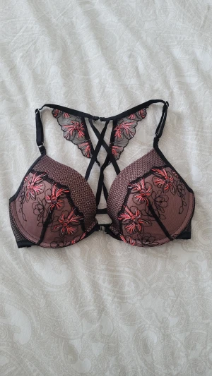 Svart behå med blommig spets - Säljer en svart behå från hunkemöller med blommig spets i rött och rosa. Toppen har snygga snörningsdetaljer framtill och meshpartier med broderade blommor. Axelbanden är justerbara och ryggen har coola korsade band. Perfekt för dig som gillar statement-plagg!