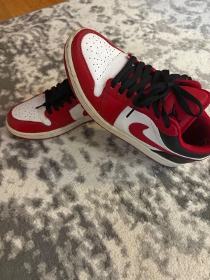 Nike Air Jordan 1 Low Chicago - Säljer ett par Nike Air Jordan 1 Low Chicago med klassisk färgblockning i rött, vitt och svart. Skorna har svart snörning, perforerad tå och rund tå. Ikonisk Air Jordan-logga på hälen och Swoosh på sidan. Tillverkade i skinn med låg siluett och platt sula. Köpa från footlocker 