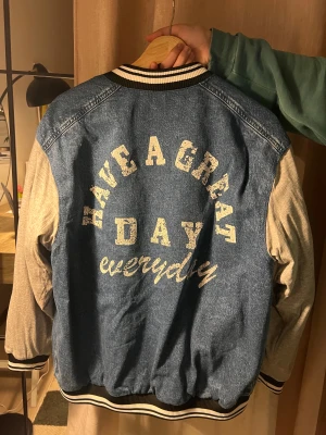 Blå jeans bomberjacka med tryck - Oversize bomberjacka i blått jeanstyg med grå ärmar i bomull. Perfekt för dig som gillar streetstyle. 