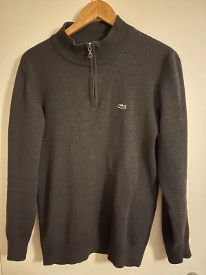 Mörkgrå stickad polotröja Lacoste - Snygg mörkgrå stickad polotröja från Lacoste med half zip och klassisk krokodillogga på bröstet. Tröjan har långärmad passform och ribbad krage som ger en clean och stilren look. Perfekt för dig som gillar enkel och tidlös design.