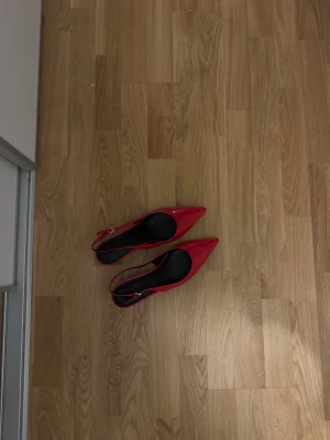 Röda pumps med spetsig tå och klack - Snygga röda pumps med spetsig tå och smal klack. Skorna har en elegant rem runt hälen med silverfärgat spänne. Ytan är blank och ger en riktigt cool look, perfekt för dig som gillar att sticka ut. Svart innersula och stilren design.