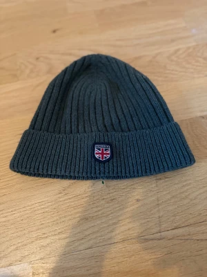 Mörkgrå ribbad mössa Morris - Snygg mörkgrå ribbstickad mössa från Morris, modell Herman Beanie. Klassisk form med uppvikt kant och Morris-emblem framtill. Perfekt för kalla dagar och enkel att matcha med din jacka. Materialet är mjukt och värmande ull.