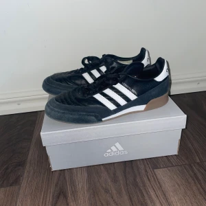 Nypris: 1750kr Futsal Adidas Mundial Goal svart/vit 39 1/3 - Adidas Mundial Goal sneakers i svart och vitt med klassiska tre ränder på sidan. Skorna har en rund tå, snörning och gummisula i brunt. Ovandelen är i äkta läder och mocka, med vit häl och Adidas-logga. Skorna är i mycket bra skick och har används ett fåtal gånger. Skriv till mig om du har frågor😁 NYPRIS: 1749kr. 