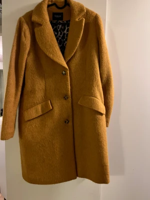 Ockragul kappa från Soaked in Luxury - Säljer en snygg ockragul kappa från Soaked in Luxury i storlek L. Jackan har klassisk krage, tre knappar framtill och två fickor med lock. Insidan har ett coolt leopardmönstrat foder som ger extra edge. Perfekt för dig som vill sticka ut i höst och vinter!