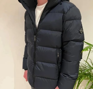 pufferjacka från J.Lindeberg - Säljer en svart pufferjacka från J.Lindeberg med huva och två sidofickor med dragkedja. Jackan har ett stilrent emblem på vänster ärm och grova, horisontella sömmar som ger en schysst look. Perfekt för kalla dagar och enkel att matcha med jeans.