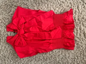 Röd blus med stor rosett från H&M - Säljer en röd råsa blus från Zara i storlek S. Blusen har en stor rosett framtill, bred smock i midjan och volangkant nedtill. Materialet är lätt och känns som bomull. Modellen är kortärmad med en snygg v-ringning och knäppning i nacken.