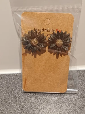 Handgjorda örhängen med blomdesign - Unika handgjorda örhängen formade som blommor i mörkbrun och guld. Blommorna har detaljerade kronblad och en guldfärgad mitt, vilket ger dem en cool och naturinspirerad look. Perfekt för dig som gillar statement-smycken med personlig touch.