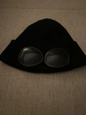 Mössa från C.P. Company - Svart ribbstickad mössa från C.P. Company med ikoniska goggle-linser framtill. Mössan är tillverkad i mjuk bomull och har en cool, urban vibe som sticker ut. Perfekt för dig som vill ha streetstyle med edge.