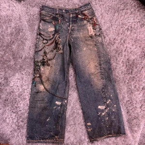 Acne Studios Graphic Jeans Baggy EUR 46 - Unika blå jeans från Acne Studios med custom fake tryck detaljer som kedjor, nyckelringar och färgstänk. Raka ben, tapered baggy passform och råa benslut. Jeans i bomull med coola slitningar och grym streetkänsla. Perfekta för dig som vill sticka ut. Väldigt bra skick.