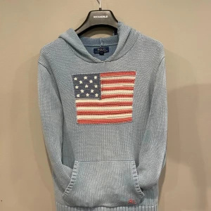 Ljusblå stickad hoodie Polo Ralph Lauren - Säljer en ljusblå stickad hoodie från Polo Ralph Lauren med amerikansk flagga broderad på bröstet. Tröjan har huva, känguruficka och RL-broderi nertill. Perfekt för dig som gillar klassisk och avslappnad stil.