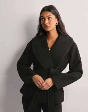 Svart kappa från Nelly - Säljer en svart kappa med bred krage och omlott-design. Jackan har ett matchande bälte i midjan och långa ärmar. Stilen är clean och modern, perfekt för dig som gillar minimalistiska vibes. Materialet ser ut att vara mjukt och lite tjockare, typ ull eller polyester.