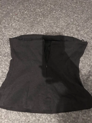 Mörkgrå tubtopp med dragkedja - Säljer en stilren mörkgrå tubtopp med dragkedja framtill. Toppen är axelbandslös och har en snygg, figurnära passform som ger en clean look. Materialet känns som bomull eller polyester och toppen är helt enfärgad utan mönster.