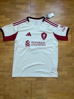 Liverpool vit matchtröja Isak #9 Adidas M - Liverpool FC vit fotbollströja från Adidas i slim fit, med röda detaljer och svarta inslag på kragen. Tryck med Isak och nummer 9 på ryggen, samt klubbmärke och sponsorlogga på bröstet. Tillverkad i lätt polyester med Aeroready-teknologi.